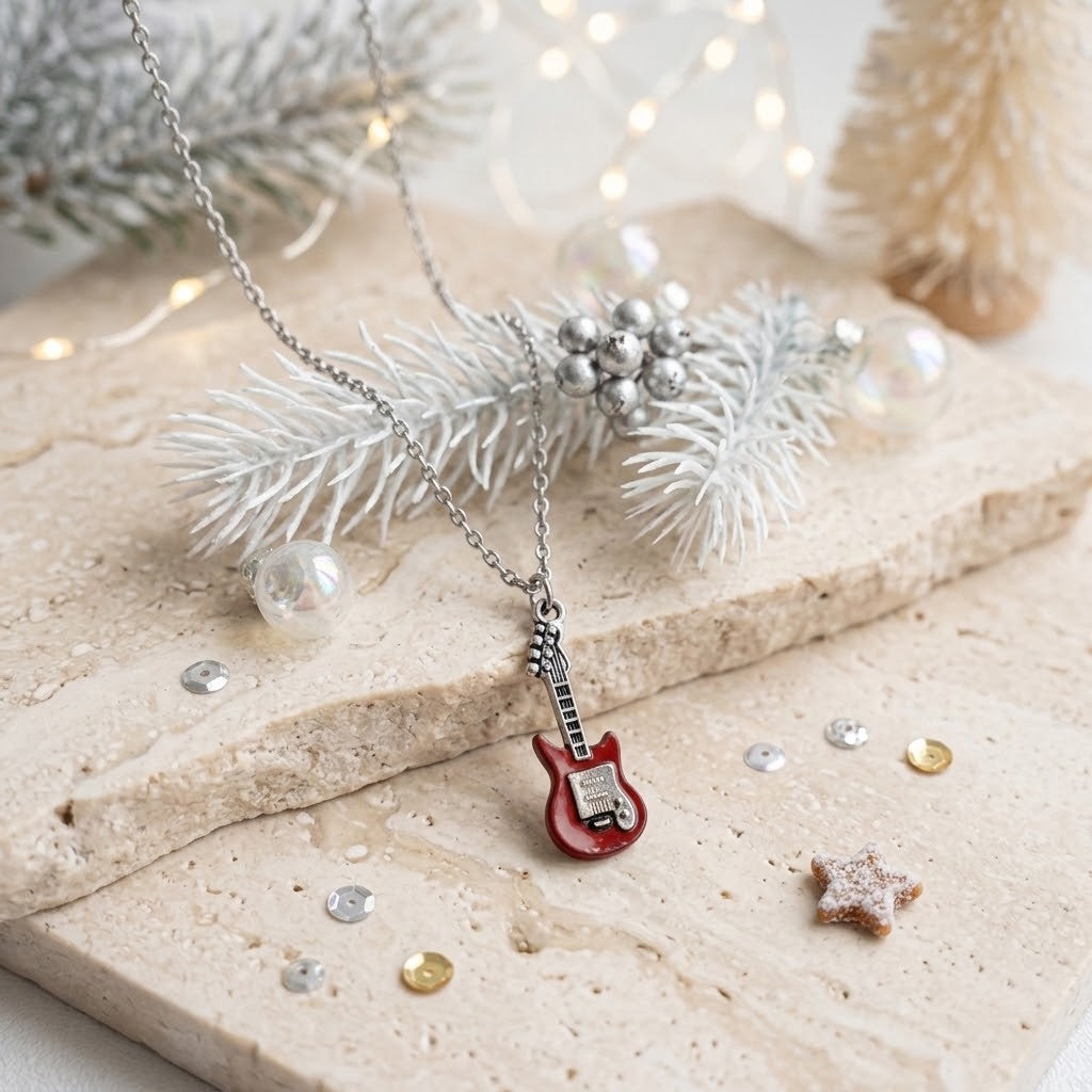 A silver-toned necklace with a small red electric guitar pendant, styled on a beige stone slab with frosted pine branches, silver beads, and warm fairy lights in the background.