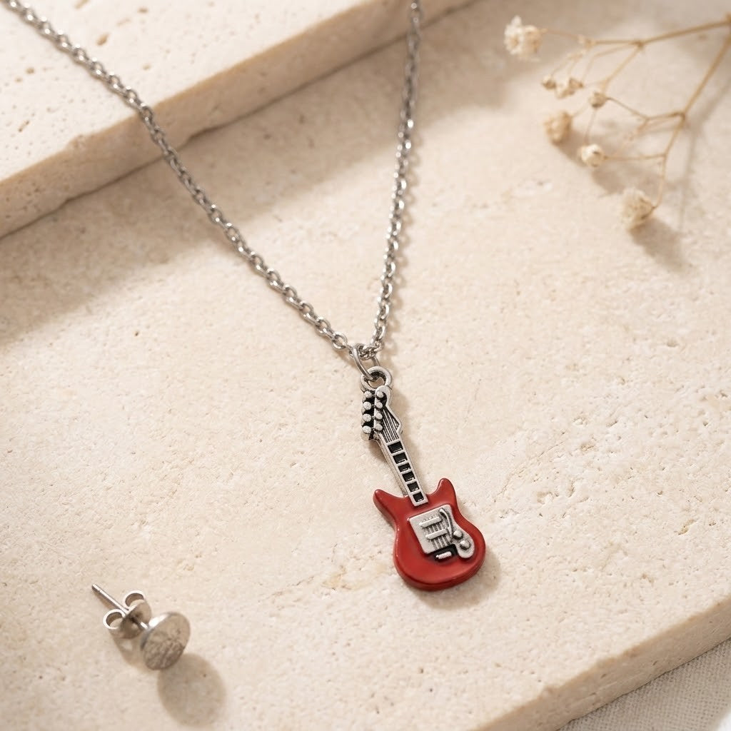A silver chain necklace with a red electric guitar pendant, displayed on a textured beige stone surface next to a matching silver stud earring.