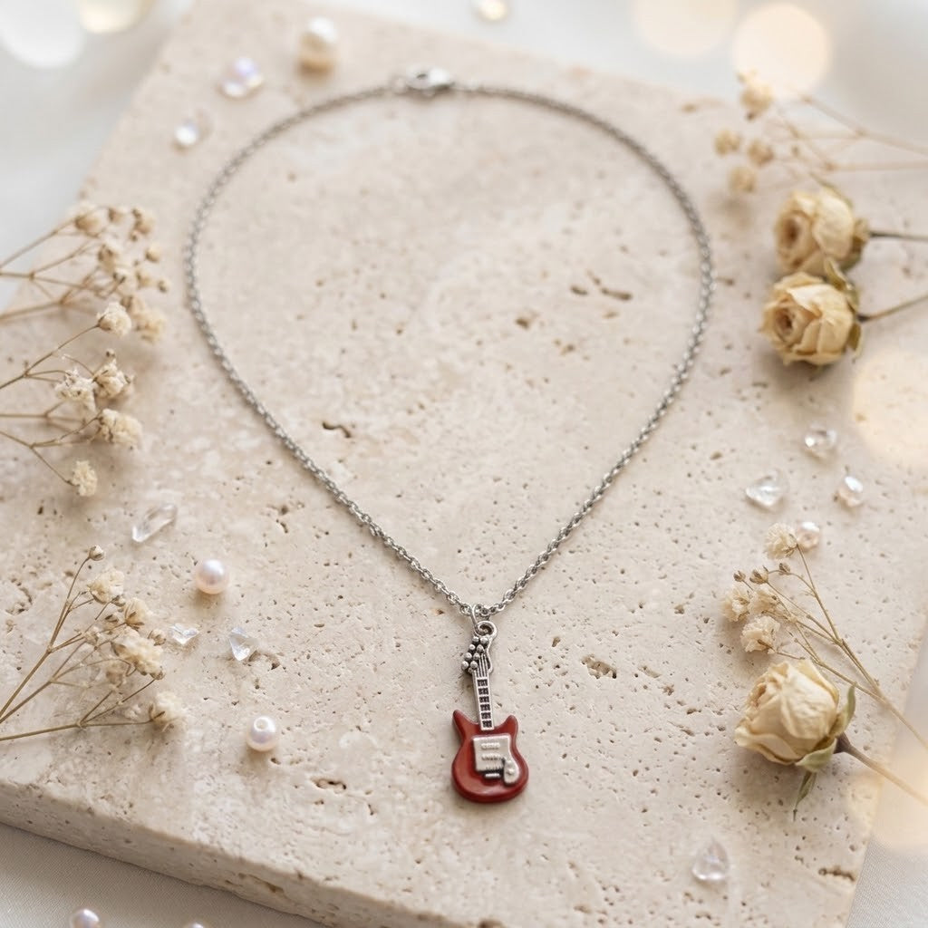 A silver chain necklace with a small red electric guitar pendant, styled on a textured beige stone slab with dried flowers and small pearls.