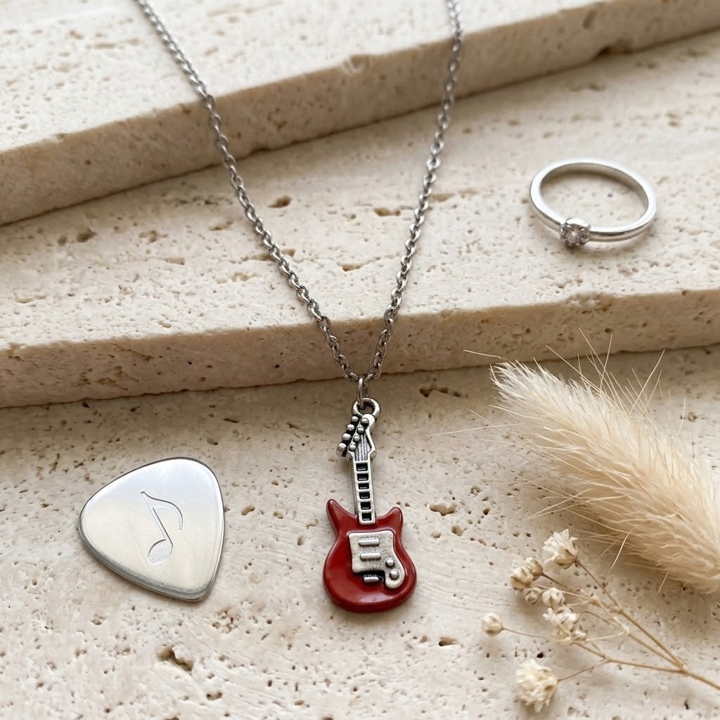 A silver necklace with a red electric guitar pendant is displayed on a textured beige stone surface alongside a silver guitar pick pendant, a silver ring, and dried grass.