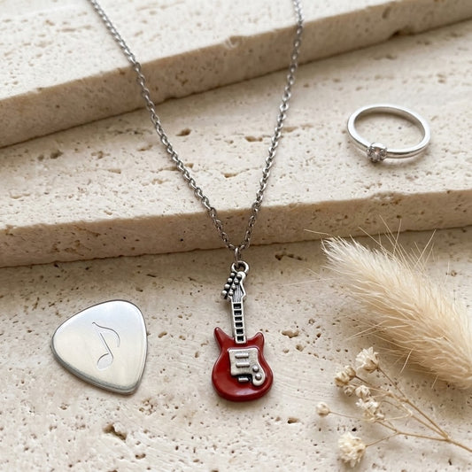 A silver necklace with a red electric guitar pendant is displayed on a textured beige stone surface alongside a silver guitar pick pendant, a silver ring, and dried grass.