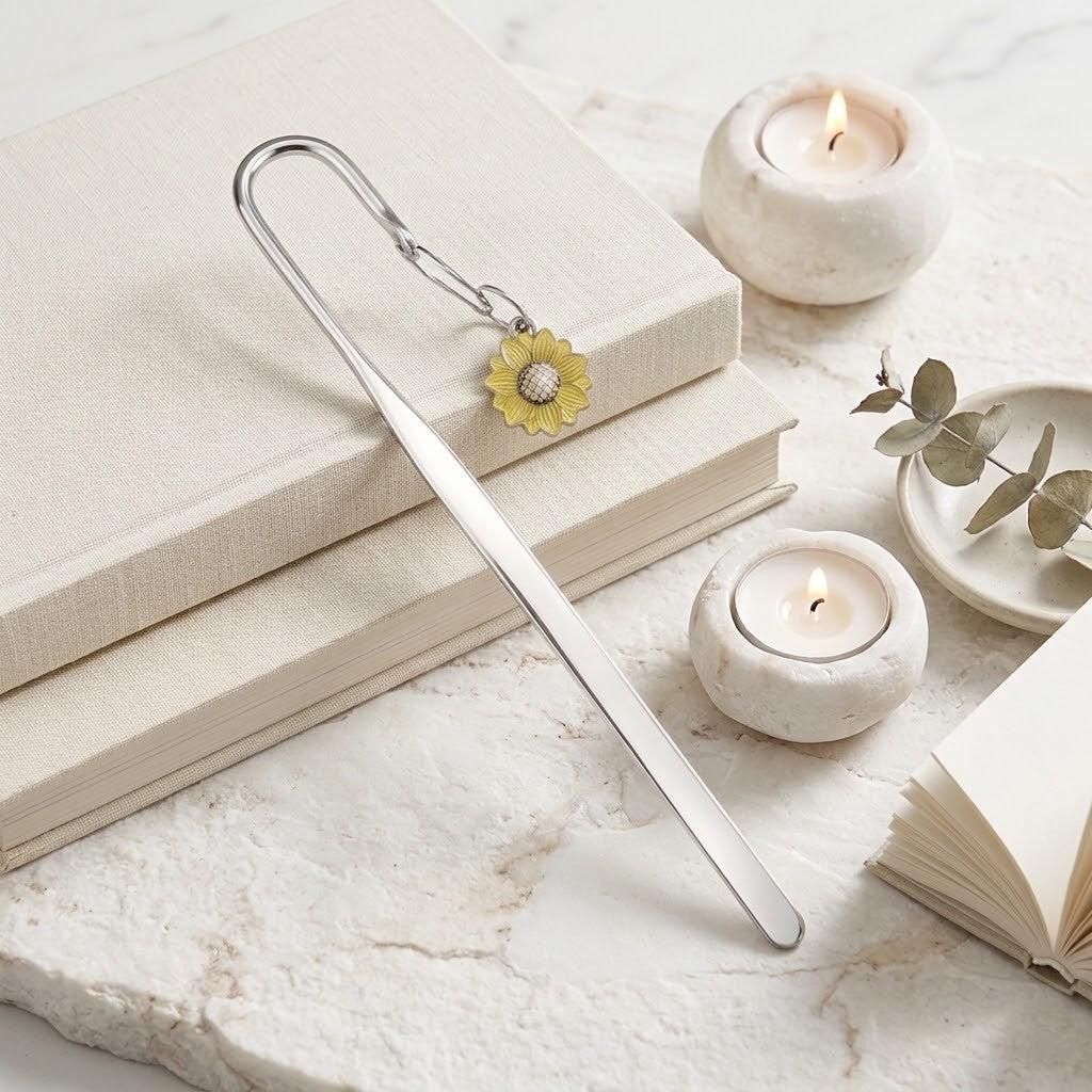 A silver metal bookmark shaped like a long needle with a small yellow flower charm, resting on stacked cream-coloured books on a light marble surface with candles and a small eucalyptus sprig nearby.