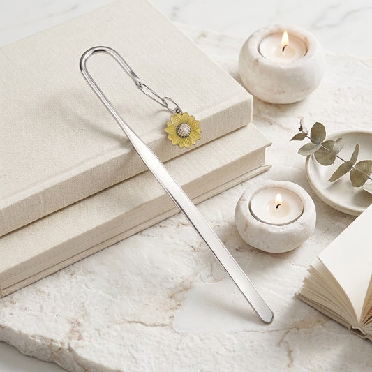 A silver metal bookmark shaped like a long needle with a small yellow flower charm, resting on stacked cream-coloured books on a light marble surface with candles and a small eucalyptus sprig nearby.