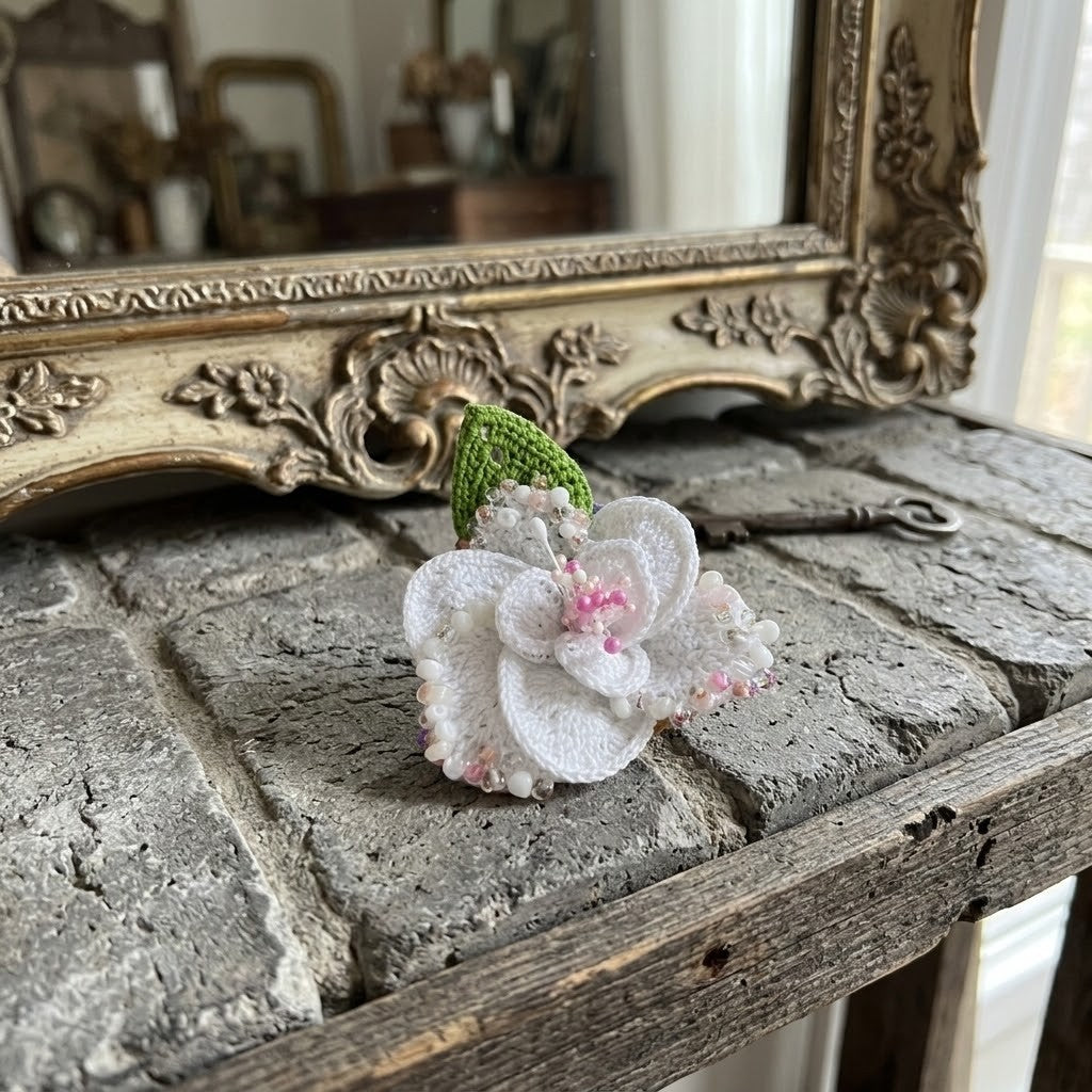 A small handmade flower-shaped brooch made of layered white fabric petals with pink stitched details and a green textured centre, resting on a rustic stone surface in front of an ornate gold mirror.