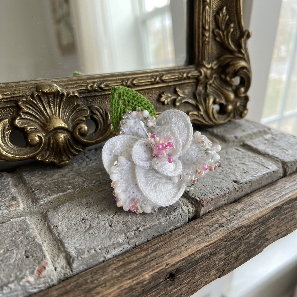 A white textured fabric flower brooch with pink bead accents and a green leaf, resting on a rustic wooden mantelpiece in front of an ornate gold mirror.
