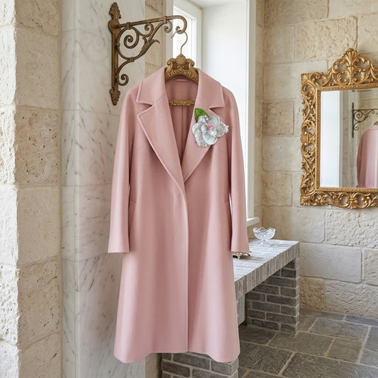 A light pink wool-blend coat with notched lapels hangs on a decorative hanger in a stone interior. A white and pink flower brooch is pinned to the left chest area.