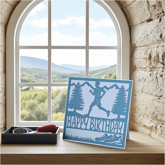 A square layered paper card with a blue silhouette of a runner, mountains, trees, and the words 'HAPPY BIRTHDAY' stands on a wooden windowsill. A small black gift box with a red item sits beside it.
