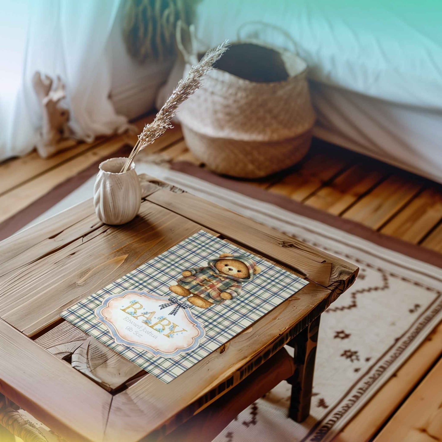 A decorative wooden table in a cosy room setting, featuring a printed card with a teddy bear illustration and the baby's name and weight. A small white vase with dried grass sits beside it, with a woven basket and soft furnishings in the background.