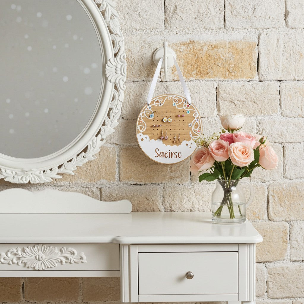A round wooden jewellery display board hanging on a white wall hook above a white vanity table, with a vase of pink roses beside it. The board has a lace cutout design, earring holes in the centre, and personalised with a name.