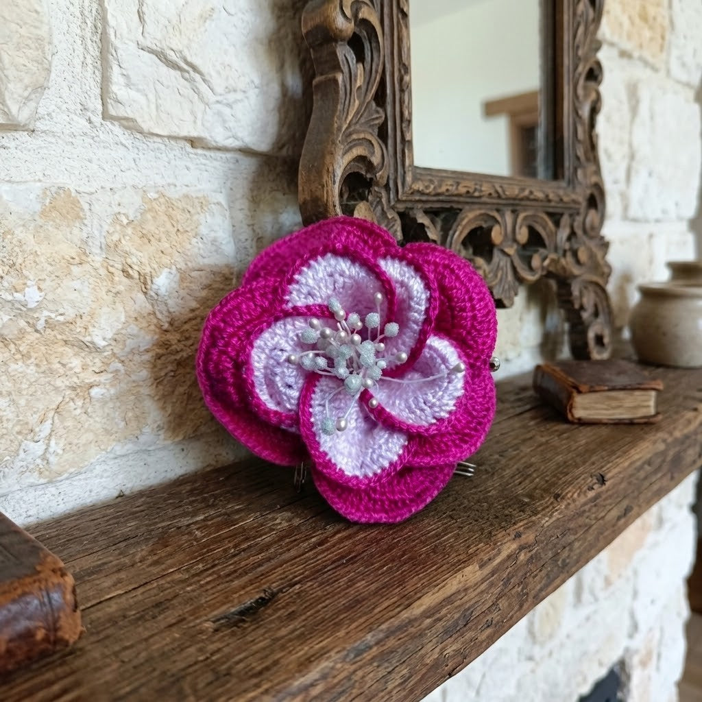A crocheted flower brooch in vibrant magenta pink and pale lavender, displayed on a rustic wooden mantelpiece against a stone wall background.