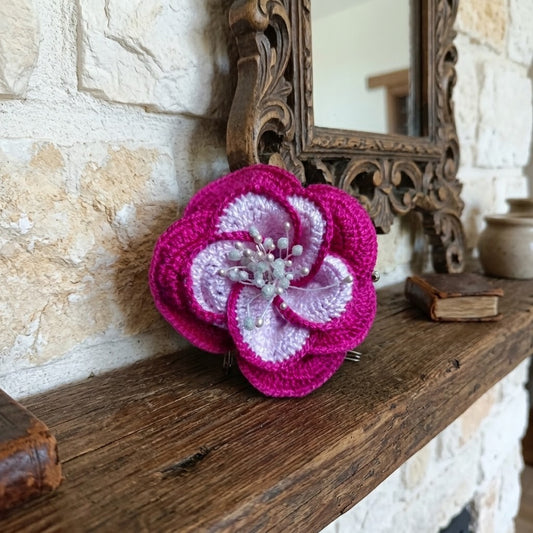 A crocheted flower brooch in vibrant magenta pink and pale lavender, displayed on a rustic wooden mantelpiece against a stone wall background.