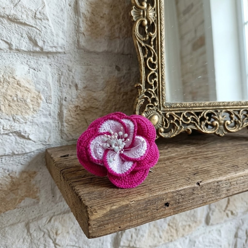 A bright pink and white crocheted flower brooch resting on a rustic wooden shelf against a light stone wall, with an ornate gold mirror partially visible.