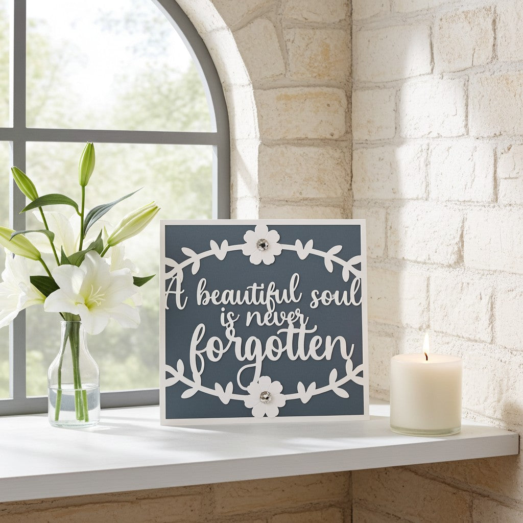 A square decorative sympathy card with the phrase “A beautiful soul is never forgotten” in white script on a dark blue-grey background, styled on a white windowsill beside white lilies in a glass vase and a lit white candle.