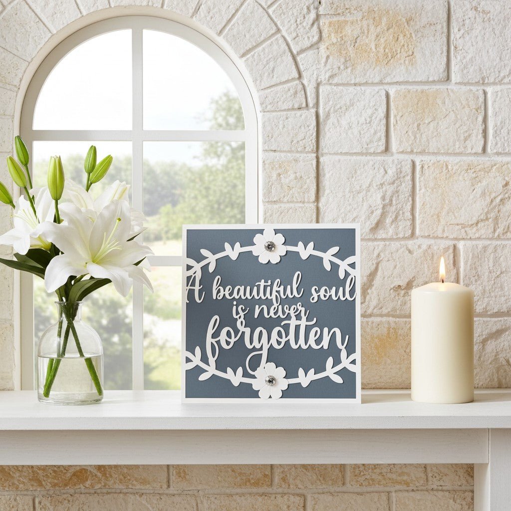 A square decorative sympathy card with the phrase “A beautiful soul is never forgotten” in white script, surrounded by a white floral wreath design on a muted blue-grey background. The sign is styled on a white mantelpiece next to a lit cream pillar candle and a glass vase of white lilies, set against a light stone brick wall and arched window.