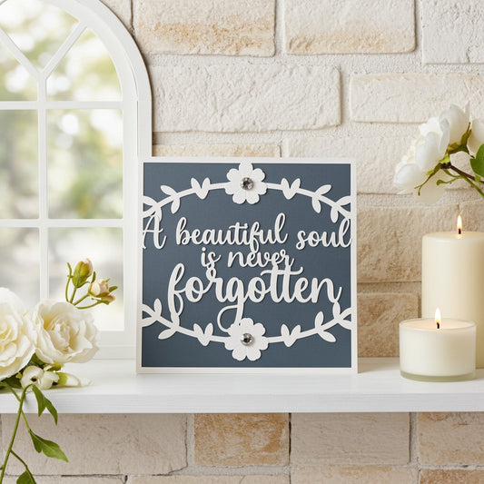 Square decorative sympathy card with the phrase “A beautiful soul is never forgotten” in white script on a dark blue-grey background, styled on a white shelf beside white flowers, candles, and a light brick wall.