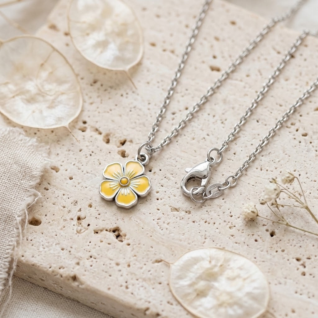 Two silver chain necklaces with flower pendants, one featuring yellow and white enamel petals and the other showing a silver lobster clasp, displayed on a textured beige stone slab with dried flowers.