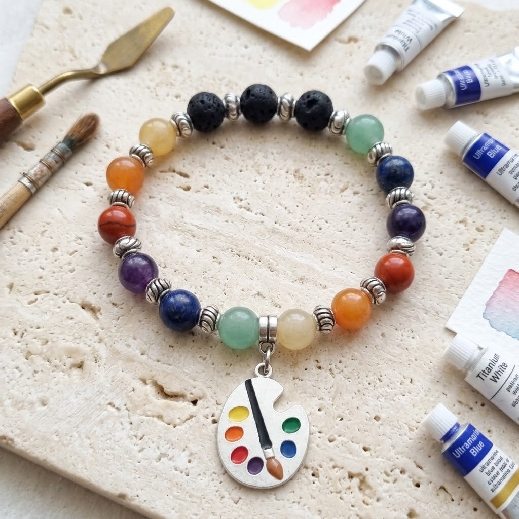 A beaded artist charm bracelet displayed on a textured stone surface, surrounded by paint tubes, a paintbrush, and a palette knife. The bracelet features a variety of colourful gemstone beads, silver spacers, and a silver paint palette charm.