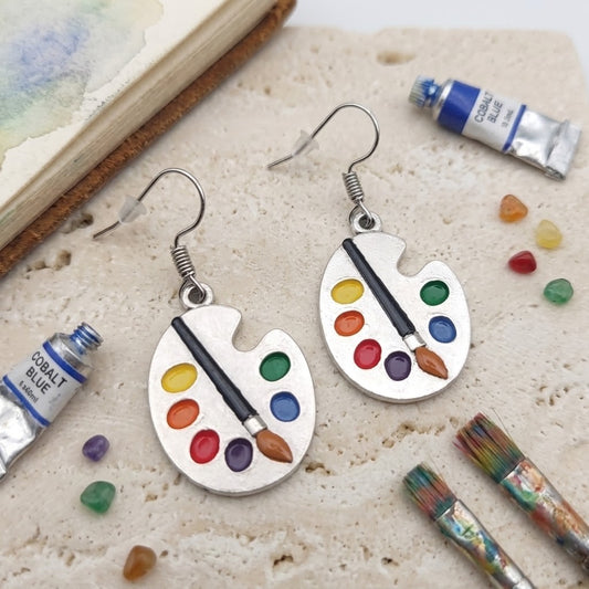 A pair of silver dangle earrings shaped like paint palettes with colourful paint droplets and a paintbrush, displayed on a textured stone surface with art props including paint tubes, brushes, and small stones.