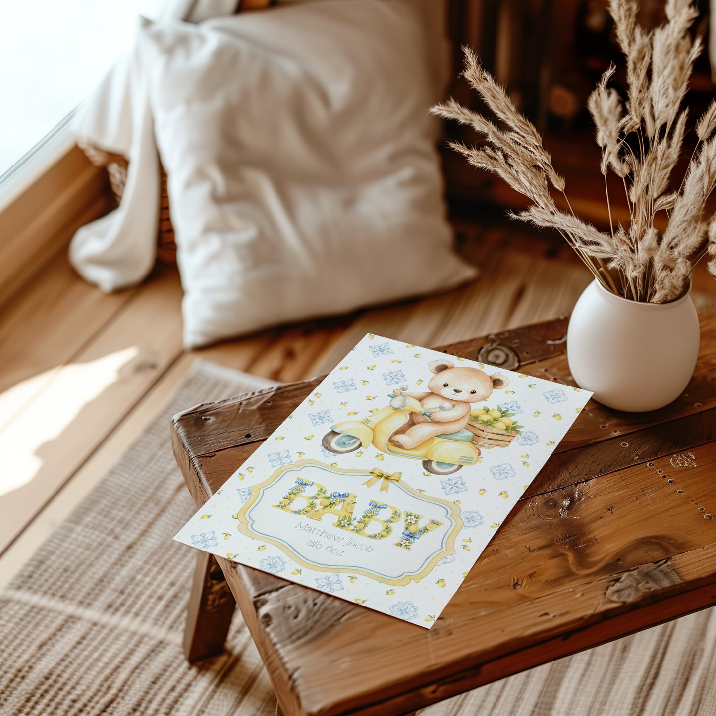 A baby card featuring a teddy bear on a yellow scooter and the word 'BABY' in blue, styled on a rustic wooden table next to a white vase with dried grasses.