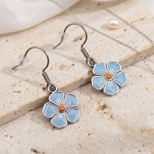 A pair of dangling flower earrings with light blue enamel petals, silver-tone metal borders, and small orange centres, displayed on a textured beige stone surface with a silver chain and a pearl nearby.