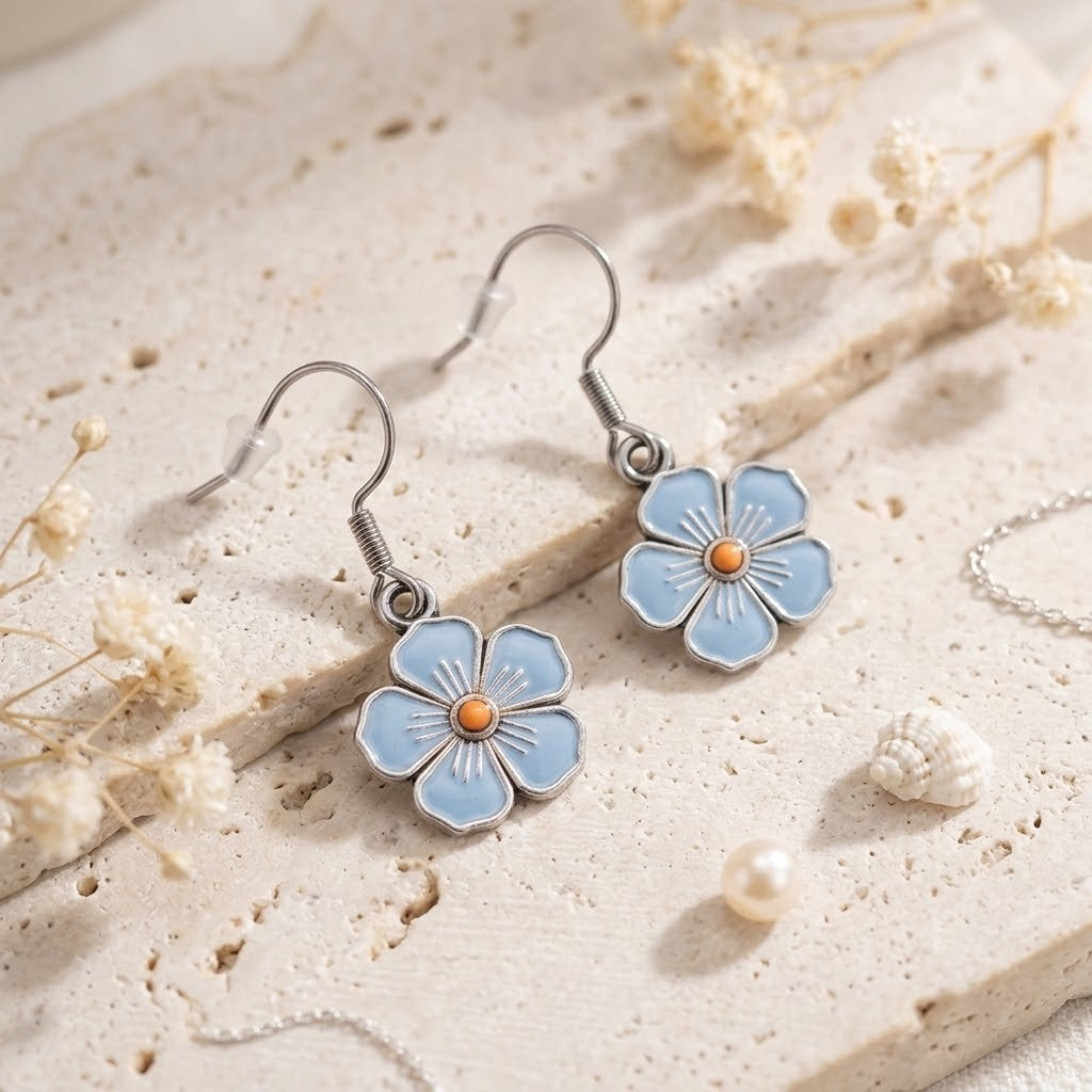 A pair of silver-tone dangle earrings with light blue flower charms and small orange centres, displayed on a textured beige stone surface with dried flowers and a pearl.