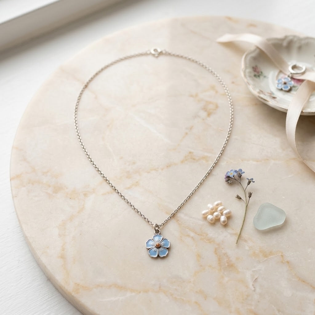 A delicate silver-tone chain necklace with a small flower pendant featuring light blue enamel and white accents, displayed on a round beige marble tray with small decorative props nearby.