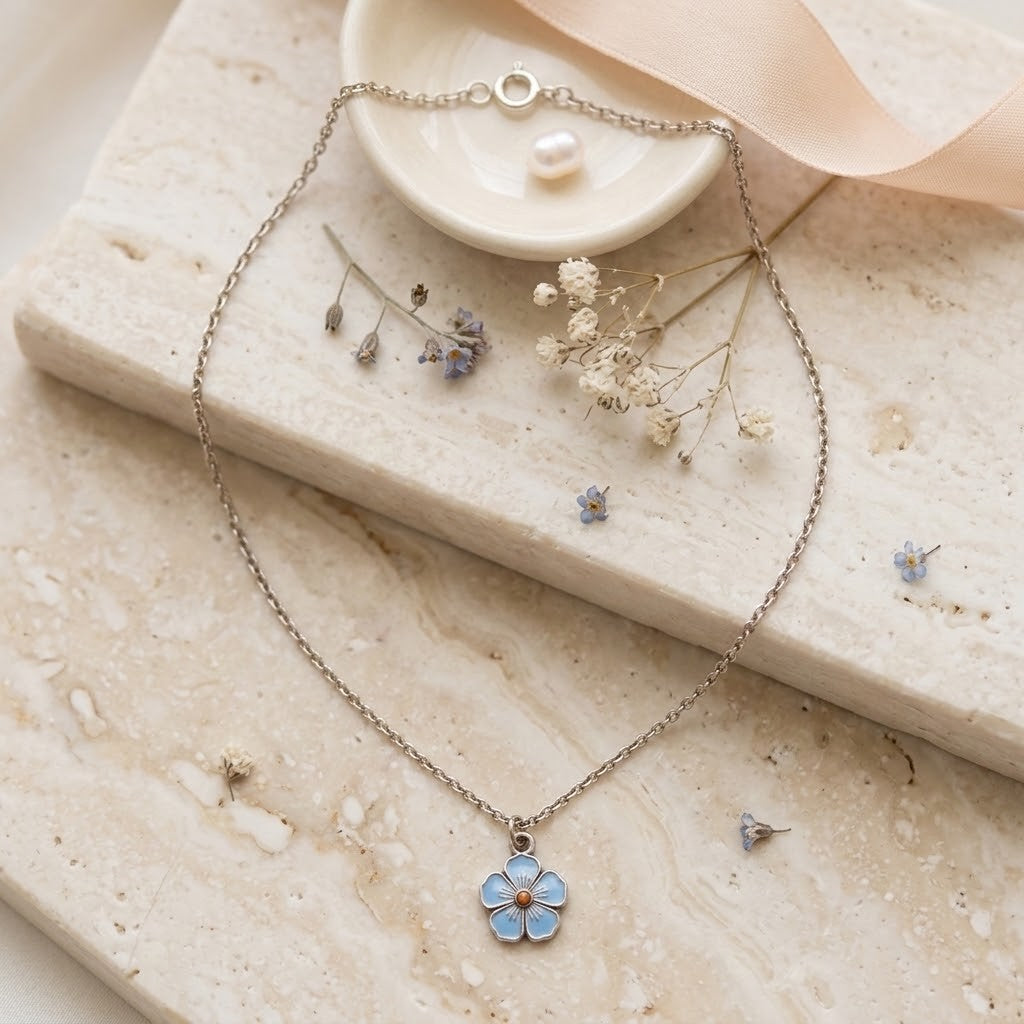 A delicate silver-tone chain necklace with a small flower pendant featuring light blue petals and a tiny gold centre, styled on a beige stone slab with dried flowers and a pale pink ribbon.