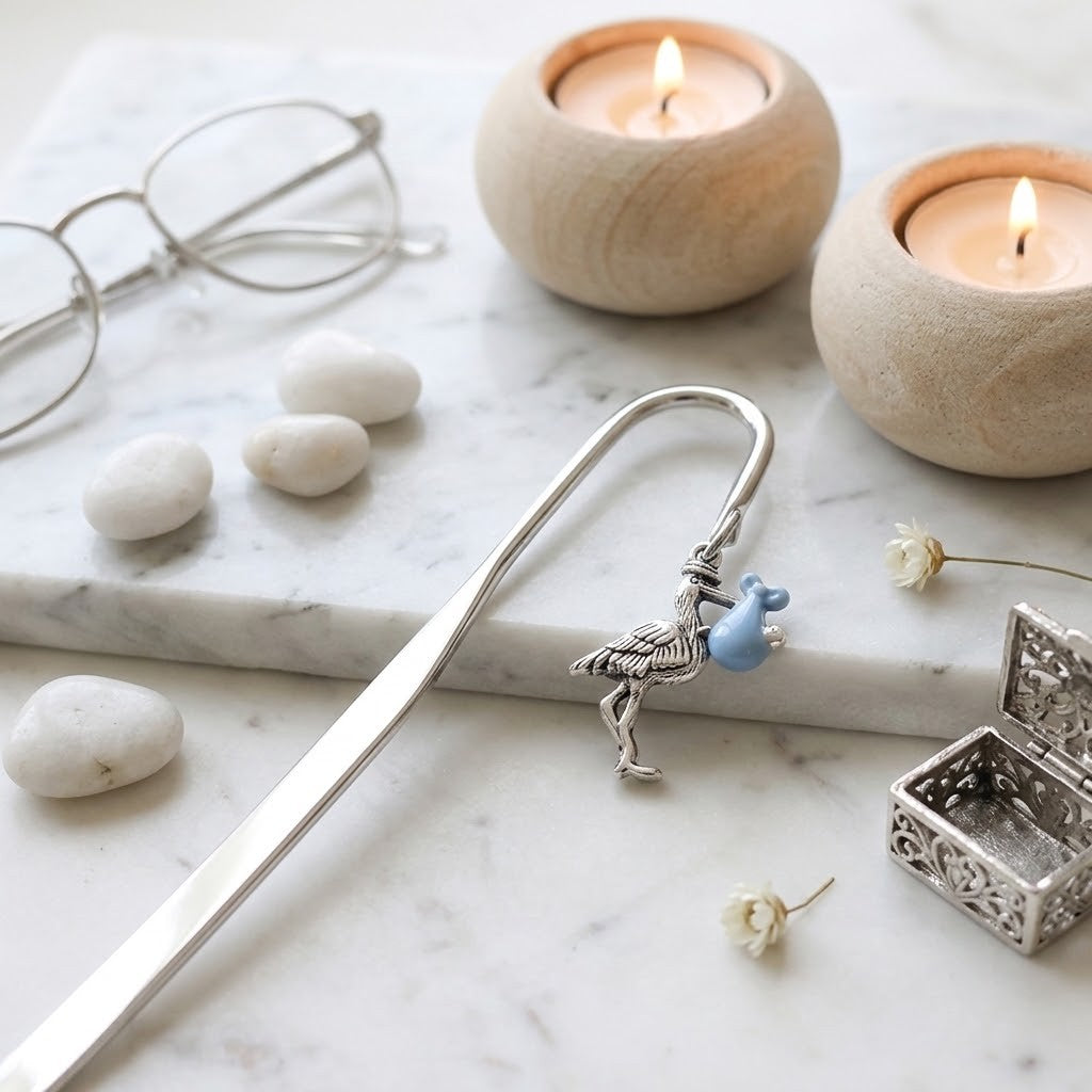 A silver metal bookmark shaped like a bird holding a small blue flower, styled on a white marble surface with glasses, white stones, two lit candles in wooden holders, and a small silver box.
