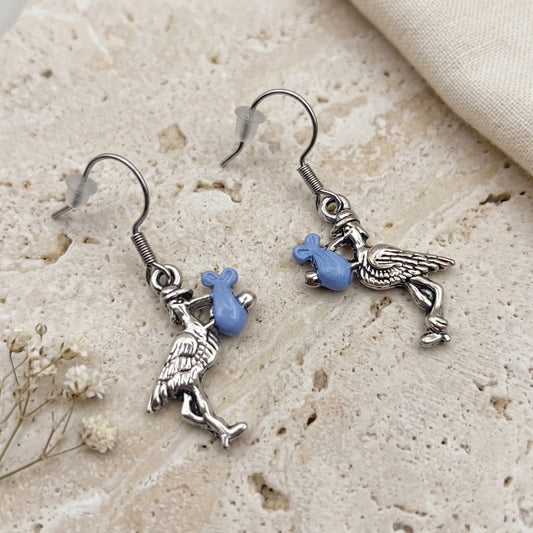 A pair of silver-toned stork earrings with small light blue bead accents, displayed on a textured beige stone surface with a folded cream fabric and dried flowers nearby.