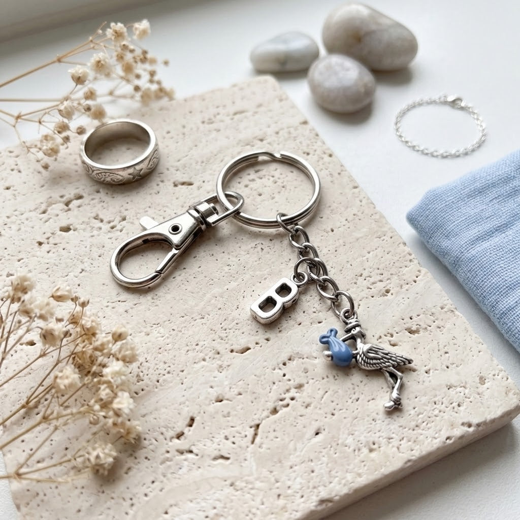 A silver-toned keychain with a lobster clasp, a hanging initial 'B' charm, and a small blue bird charm, styled on a textured stone slab with dried flowers, a ring, stones, and a folded blue cloth.