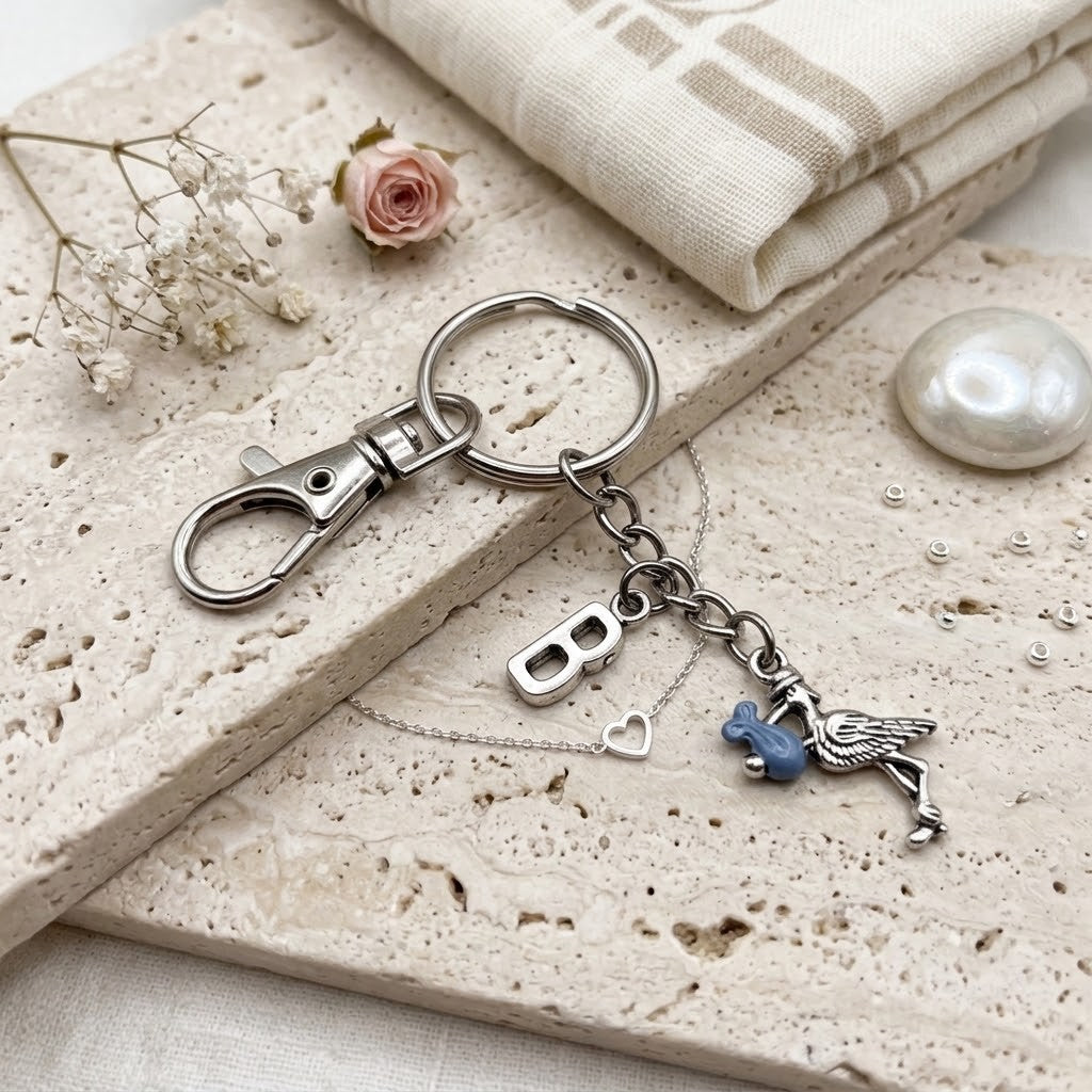 A silver-toned metal keychain with a lobster clasp, key ring, charm shaped like a stork, displayed on a textured beige stone slab with fabric and dried flowers.