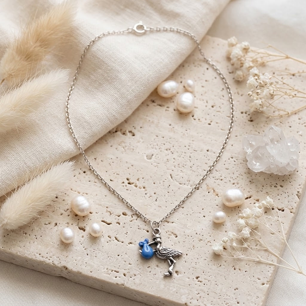 A silver-toned chain necklace with a stork pendant holding a small blue flower, styled on a textured beige stone slab with faux pearls, dried flowers, and soft cream fabric.