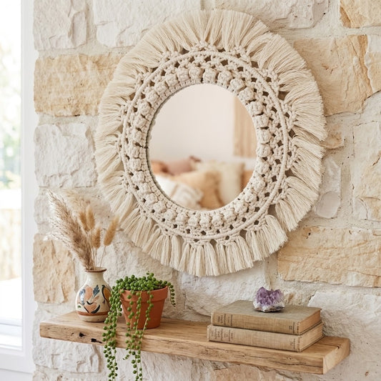 A round bohemian-style macrame mirror with a thick woven beige cord and fringe, mounted on a rustic stone wall above a wooden shelf with dried grass, a small plant, and books.