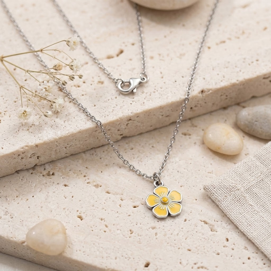 A silver-tone floral pendant necklace with a yellow and white enamel flower charm, displayed on a textured beige stone surface with small decorative stones and dried flowers.