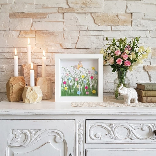 A framed art print featuring a colourful wildflower meadow with a butterfly, styled on a white wooden console table against a light stone brick wall. The frame is surrounded by decorative props including candles, a vase of pink roses, a small white elephant figurine, and books.