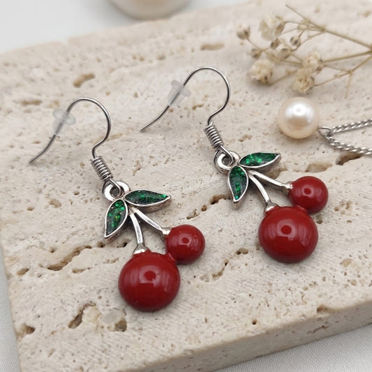 Cherry drop earrings with red bead-like cherries, green leaf charms, and silver hooks, displayed on a light stone slab with a pearl and dried flowers nearby.