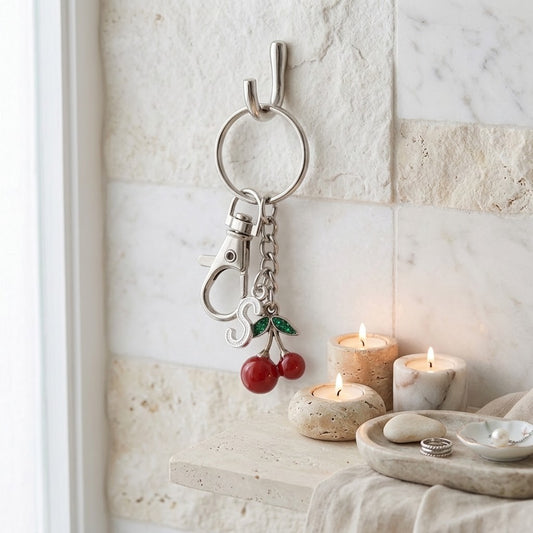A silver metal keychain with a split ring, lobster clasp, and two cherry charms hangs on a wall hook in a bathroom setting. The background features light beige stone tiles, a small shelf with lit candles.
