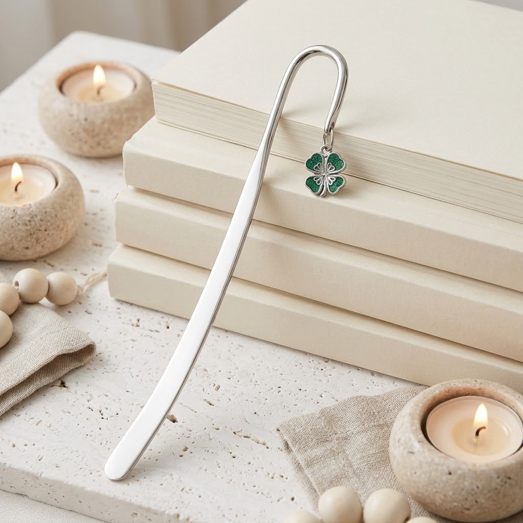 A silver metal bookmark with a green four-leaf clover charm, resting on a stack of cream-coloured books. The scene is styled with lit candles in textured holders and small wooden beads on a light stone surface.