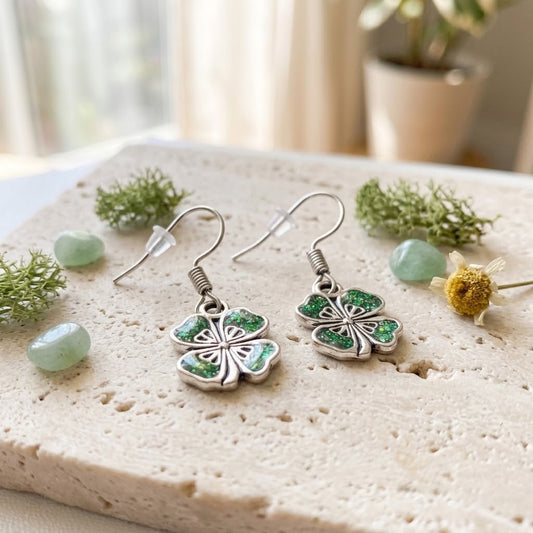 A pair of silver dangling earrings with four-leaf clover charms filled with sparkling green glitter, displayed on a textured light stone surface with small green stones and dried floral accents.