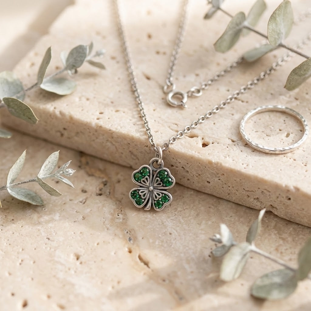 A silver-tone necklace with a four-leaf clover pendant set with green stones, displayed on a textured beige stone slab with eucalyptus leaves and a silver ring.