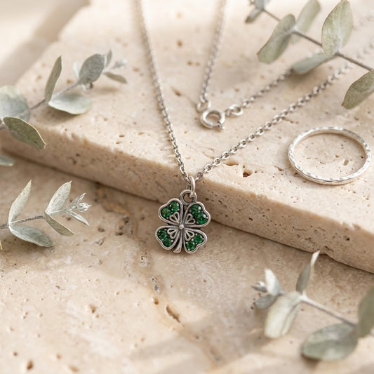 A silver-tone necklace with a four-leaf clover pendant set with green stones, displayed on a textured beige stone slab with eucalyptus leaves and a silver ring.