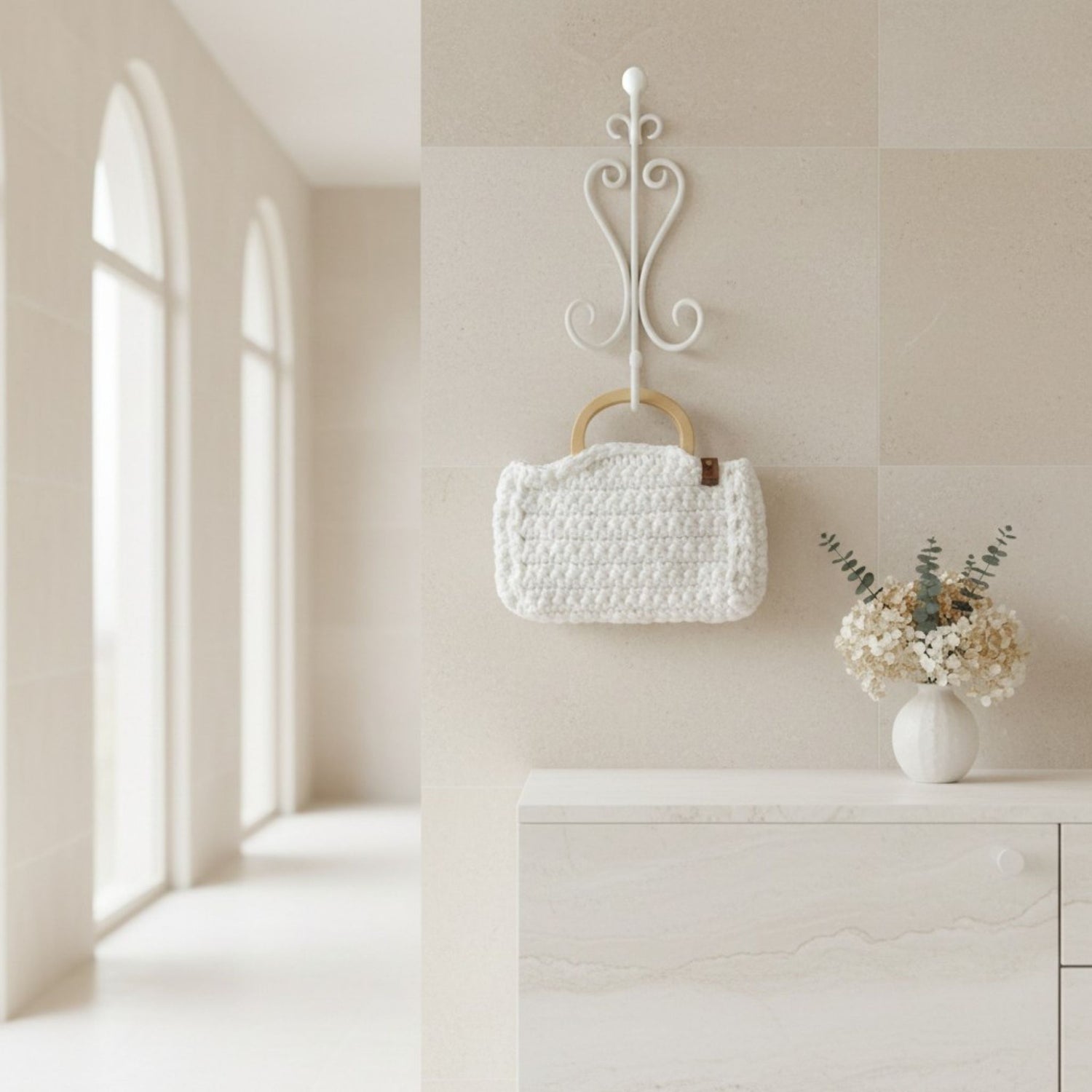 White crochet bag hung on a wall with wooden handle next to a vase of flowers.