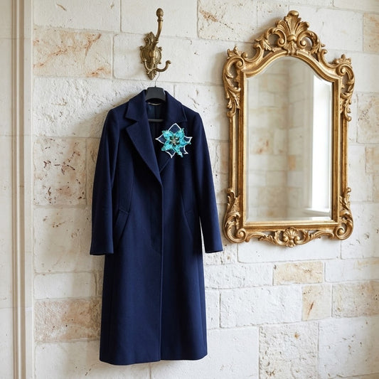 A navy blue wool coat hangs on a black hanger from an ornate brass hook on a light stone wall, next to a large gold-framed mirror. The coat features a decorative blue and turquoise embroidered star-shaped accent on the left chest.