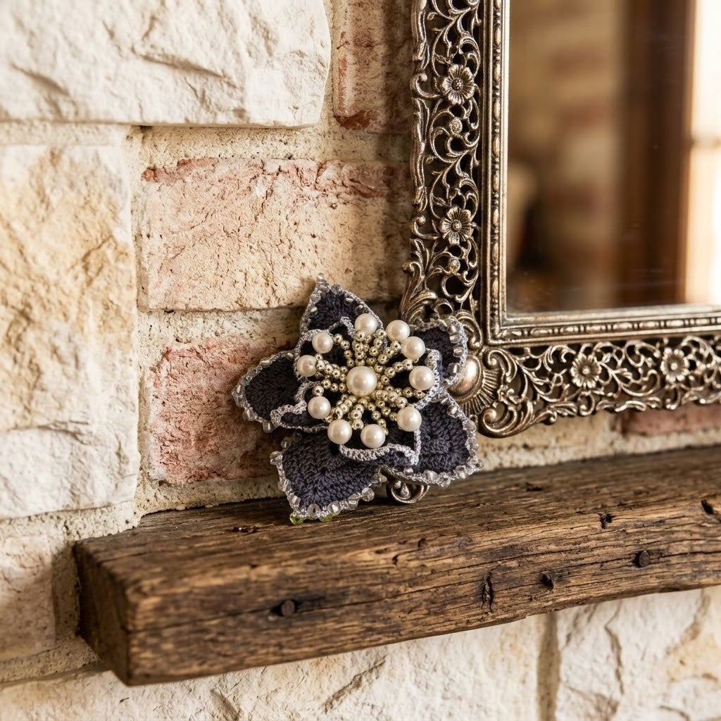 A dark grey knitted flower brooch embellished with white faux pearls and gold-toned beads, resting on a rustic wooden mantel against a stone wall next to an ornate mirror.