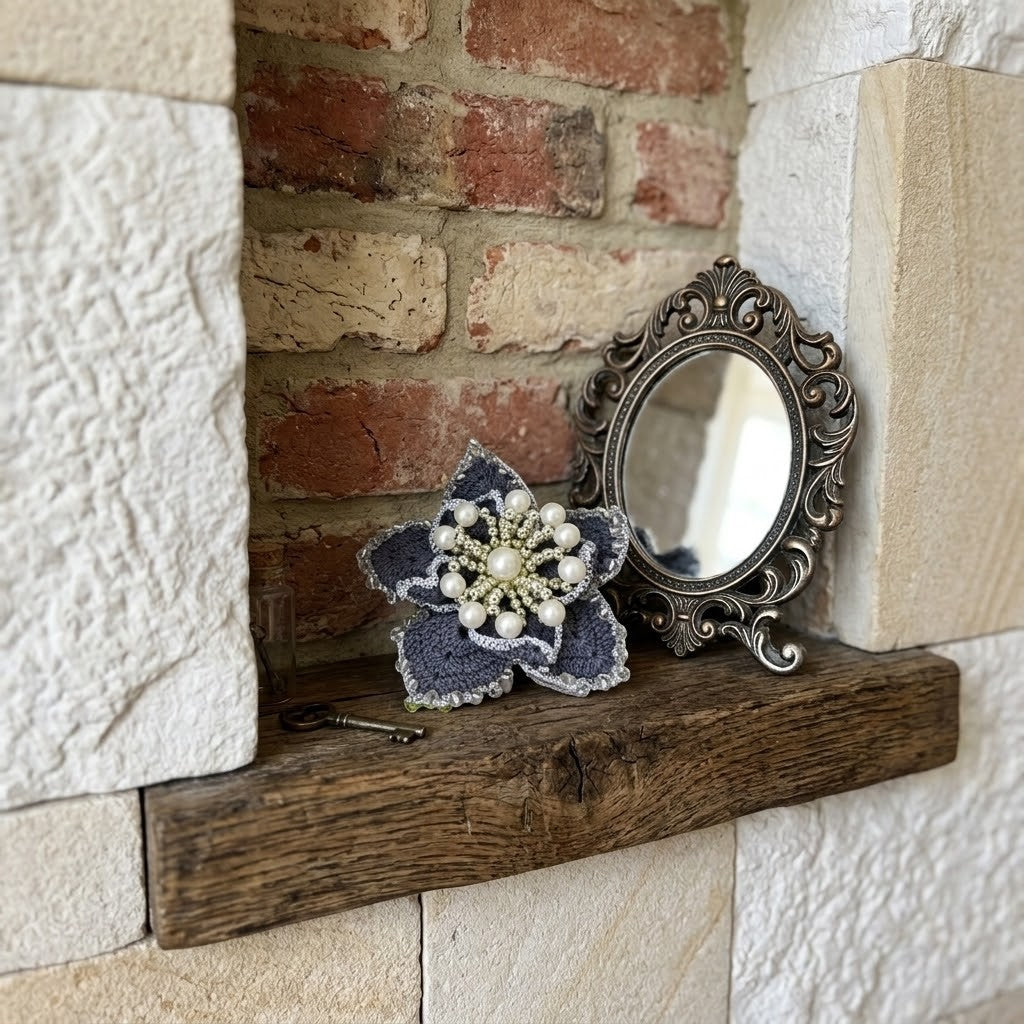 A decorative floral brooch with grey crochet petals and white faux pearls, displayed next to an ornate silver mirror on a rustic wooden shelf against a brick and stone wall.