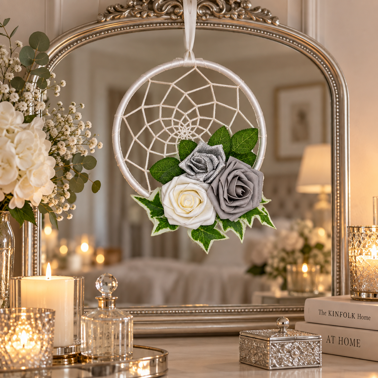 A hanging dreamcatcher ornament with white webbing, a white rose, a grey rose, and green leaves, displayed in front of an ornate mirror on a decorated tabletop with candles and flowers.