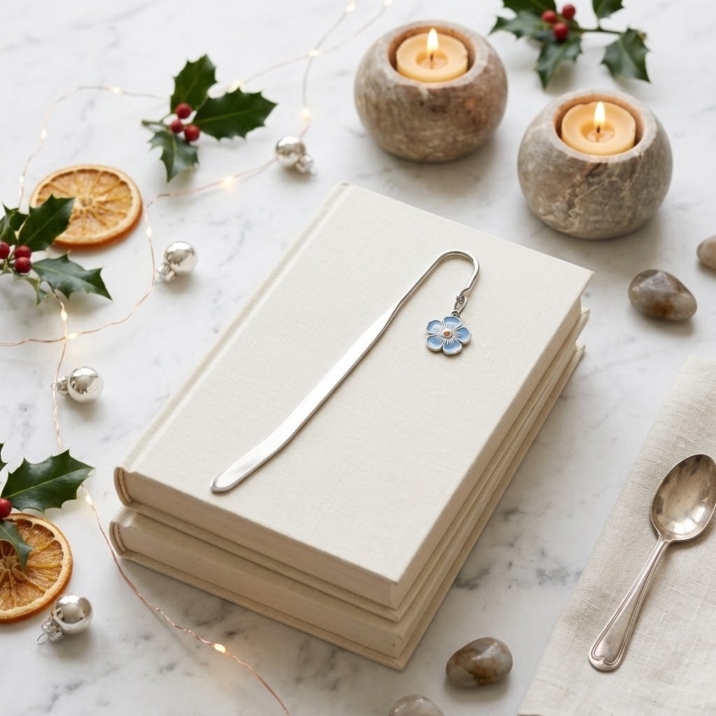 A silver metal bookmark with a small blue flower charm rests on a stack of cream-colored hardcover books. The scene is styled with a silver spoon, dried orange slices, holly leaves, silver ornaments, and two lit candles in stone holders on a white marble surface.