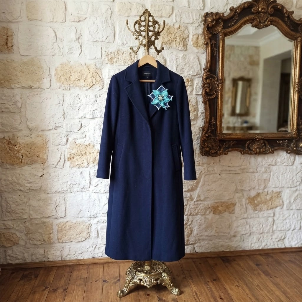 A long navy blue coat displayed on a hanger stand against a rustic stone wall with a large ornate mirror. The coat features a decorative blue and white floral brooch pinned at the chest.