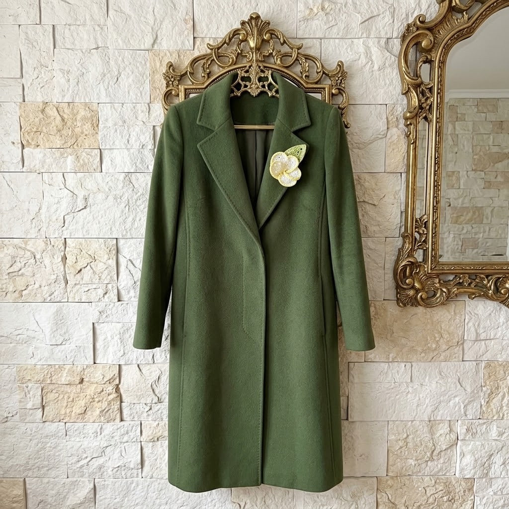 An olive green wool coat with notched lapels hangs on an ornate gold hanger against a light beige stone wall, decorated with a white flower brooch on the lapel.