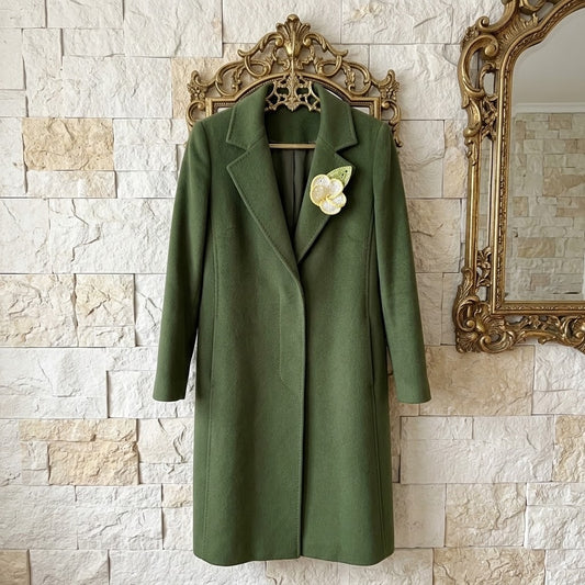 An olive green wool coat with notched lapels hangs on an ornate gold hanger against a light beige stone wall, decorated with a white flower brooch on the lapel.